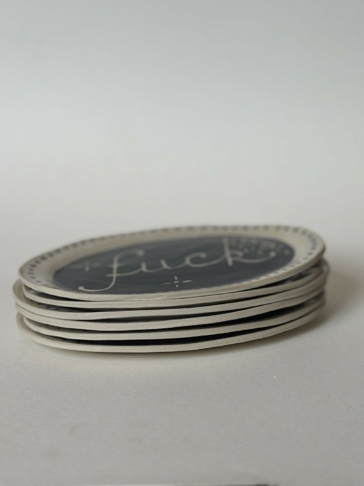 Pile d’assiettes ovales noir avec texte 'fuck' et bord à pois, empilées.