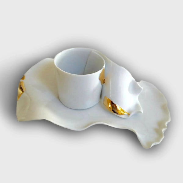 Ensemble tasse et soucoupe blanche forme irrégulière, accent ruban doré sur le côté.