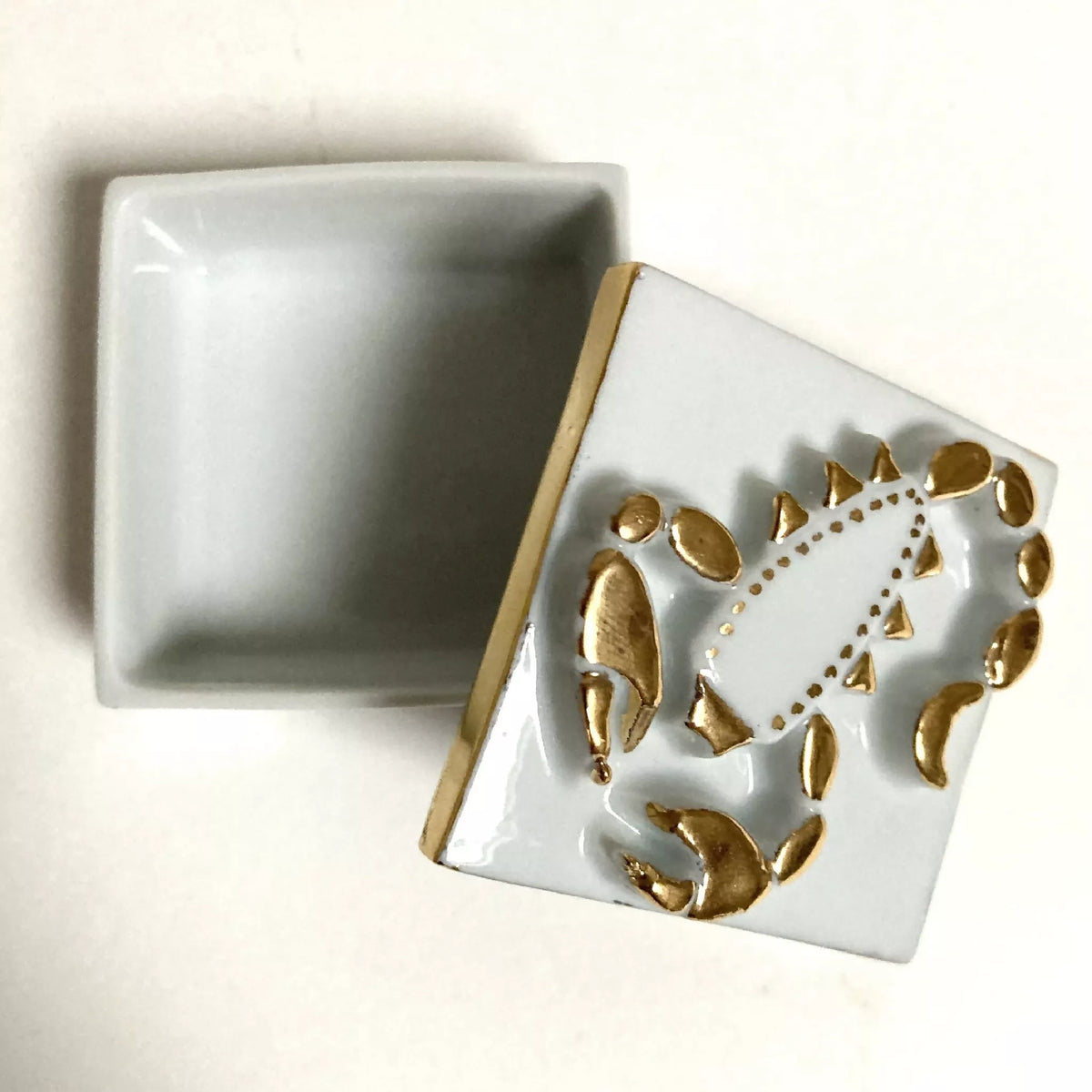Boîte à bijoux en porcelaine carrée, couvercle ouvert, motif scorpion doré sur fond clair.