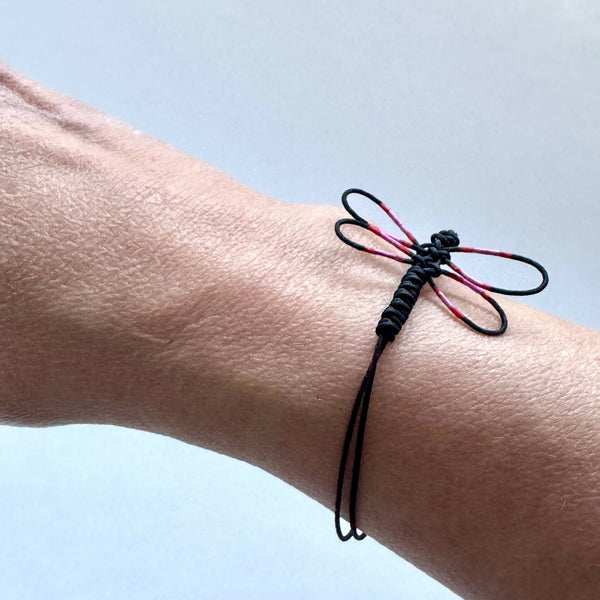 Bracelet noir en cordon tressé, libellule peinte rose et rouge près du poignet.