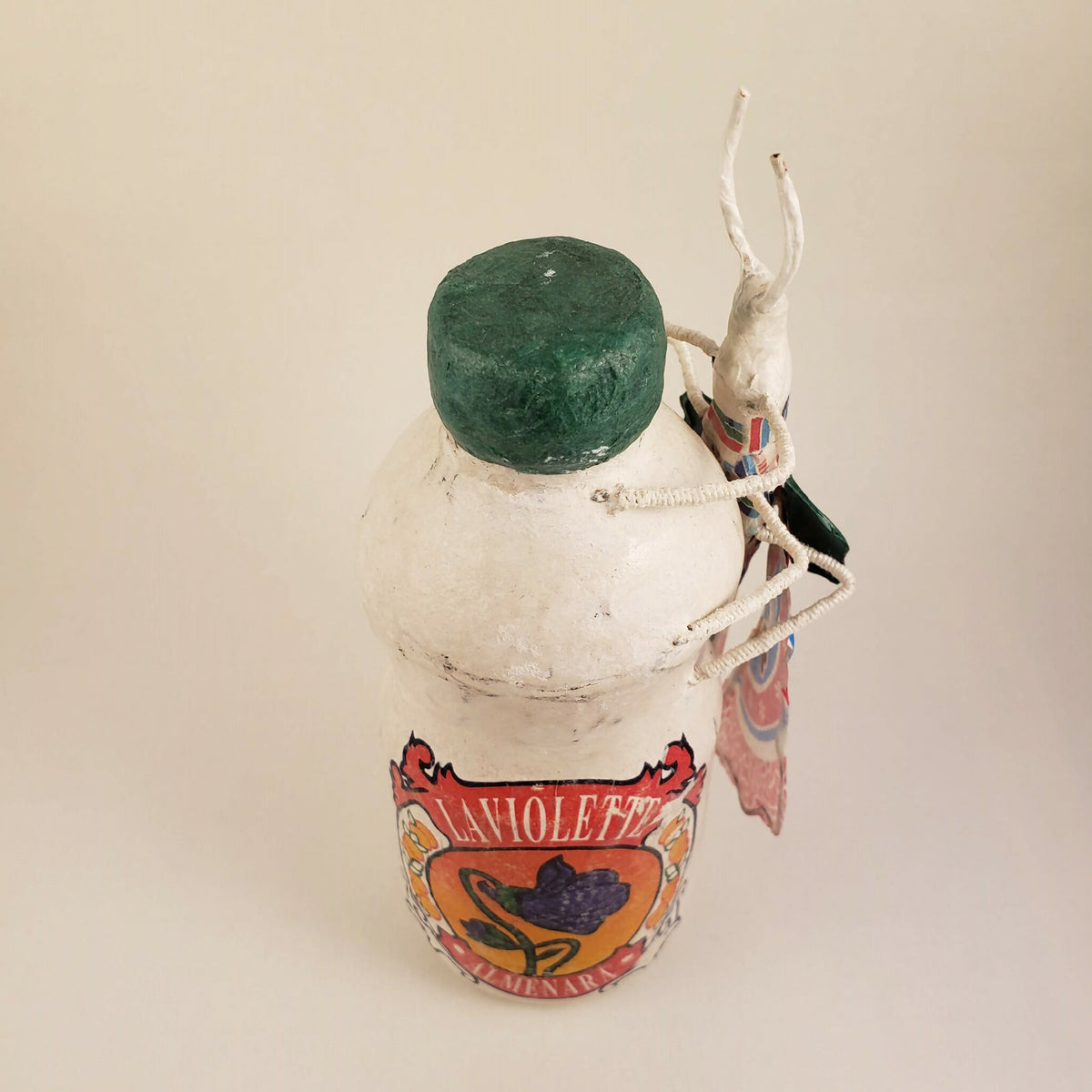Sculpture posée sur une bouteille, gros plan sur la face, capuchon vert, tablier coloré