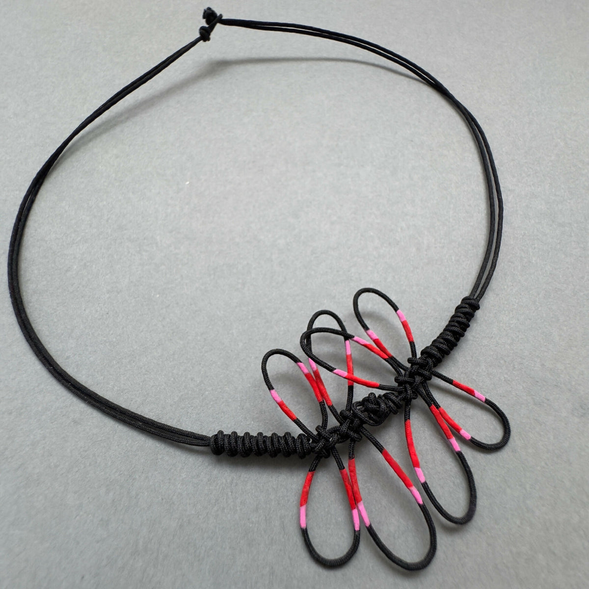 Bracelet noir en cordon tressé avec libellule rose et rouge, nœud et brins visibles.