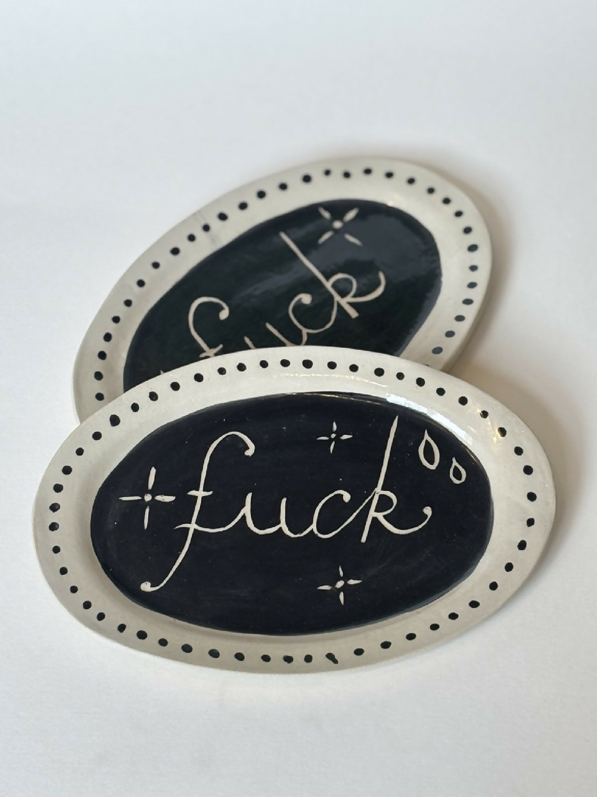 Assiettes ovales noir et beige, texte 'fuck' peint à la craie, motif pois au bord.