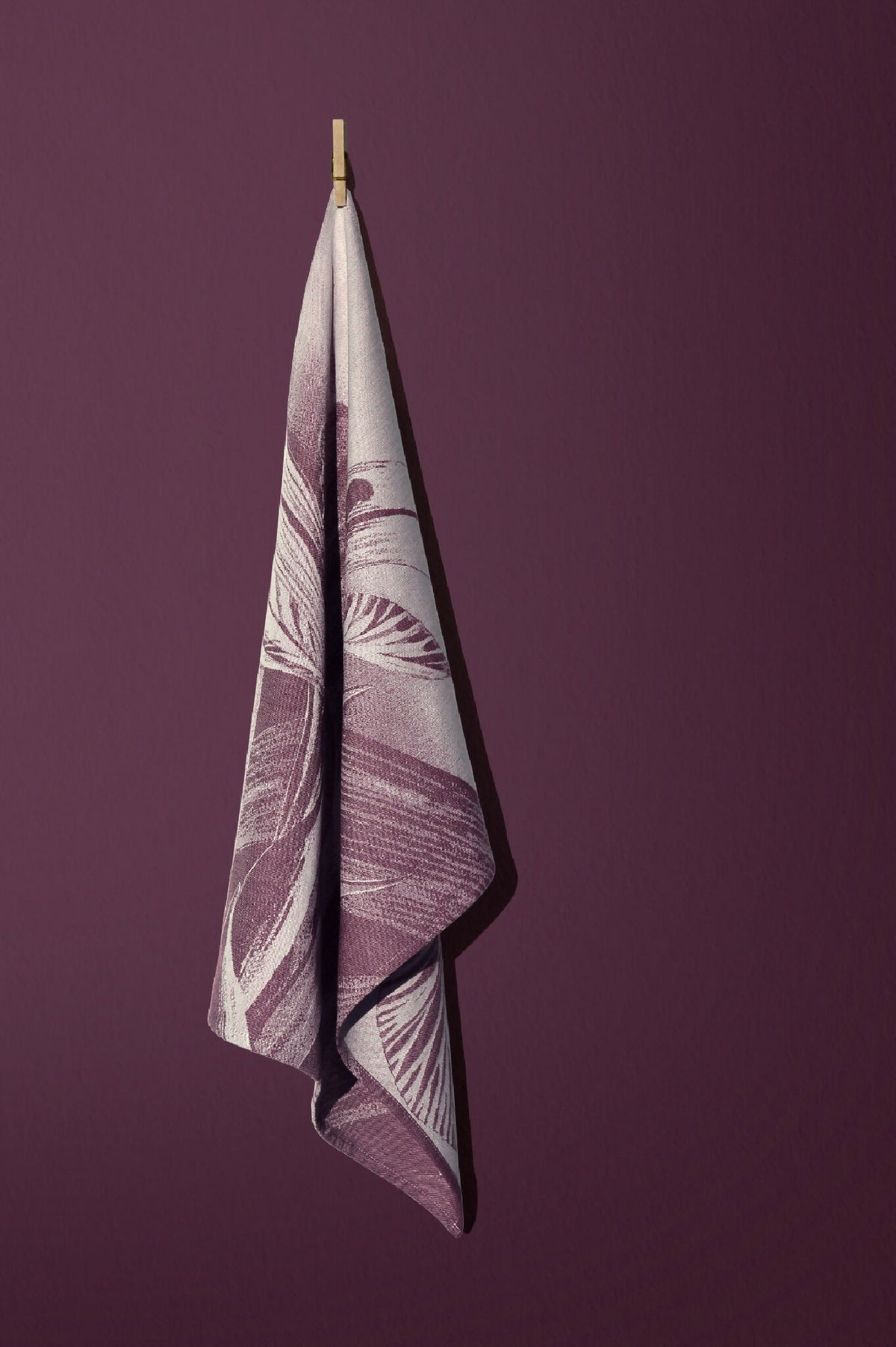 Torchon imprimé replié, motif plume et feuilles violet sur fond blanc cassé.