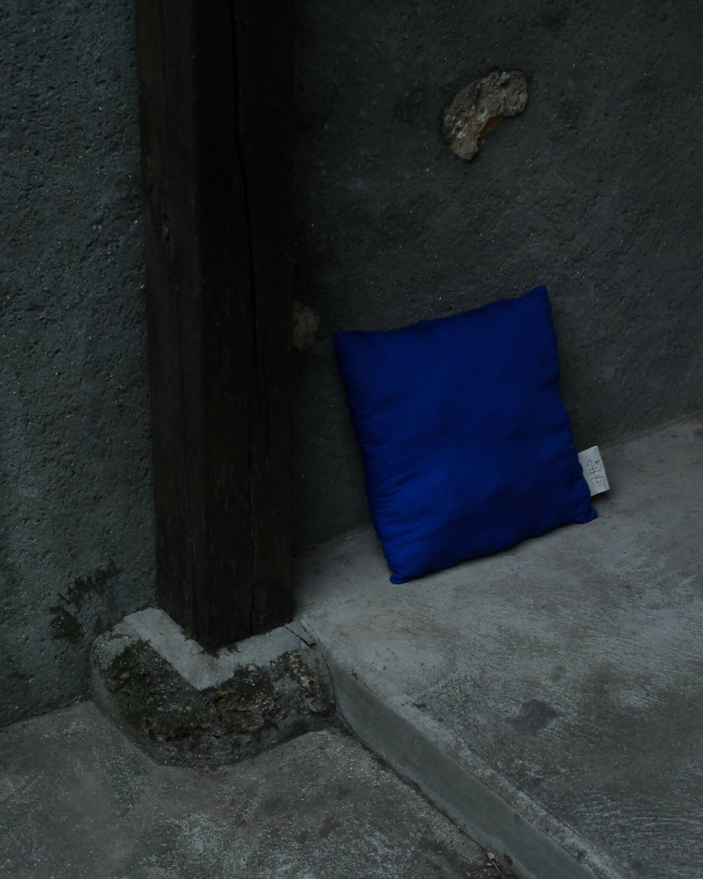 Coussin bleu carré, posé au sol, étiquette blanche visible sur le côté