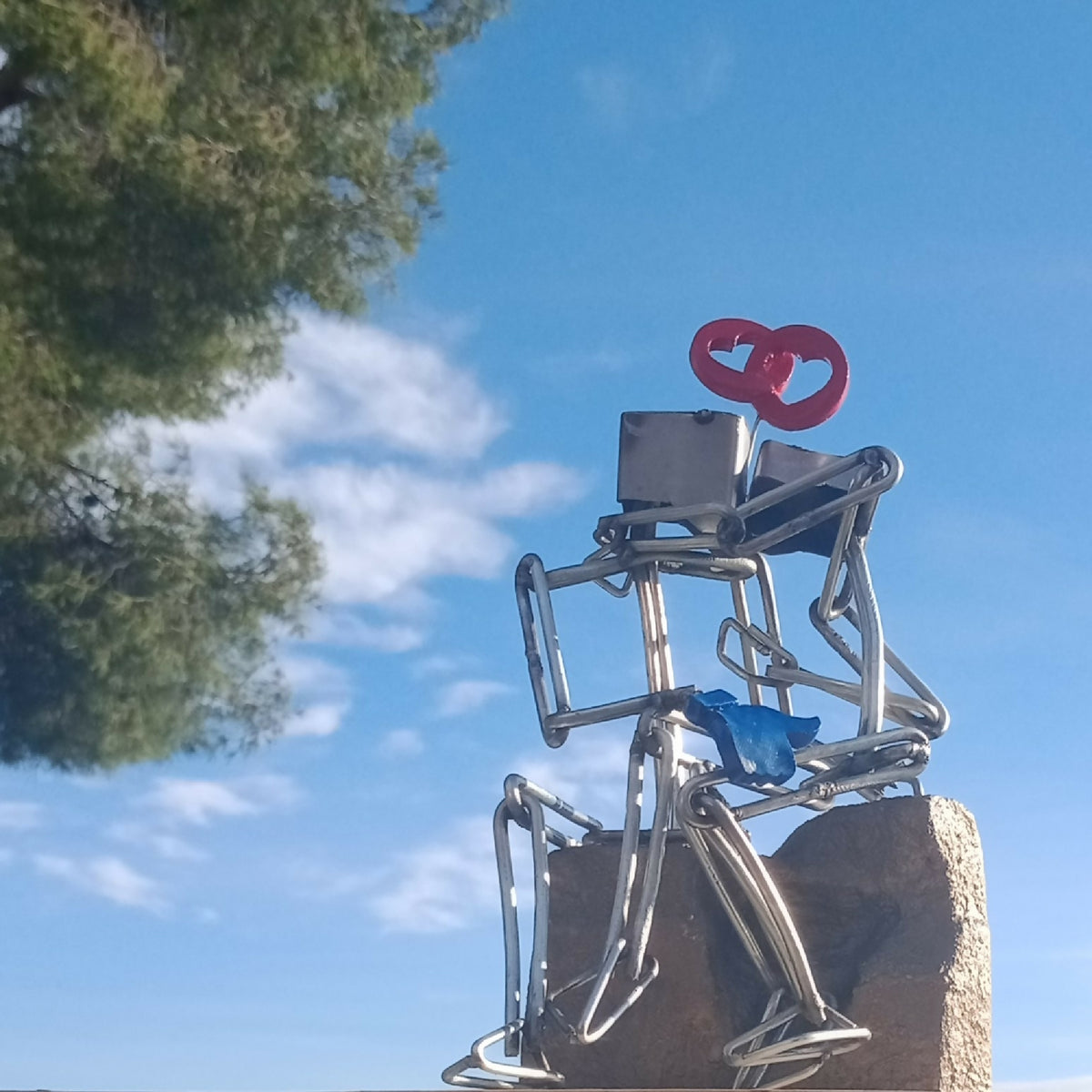 Sculpture en fils d’acier sur socle doré, couple et cœur rouge, avec vue en extérieur