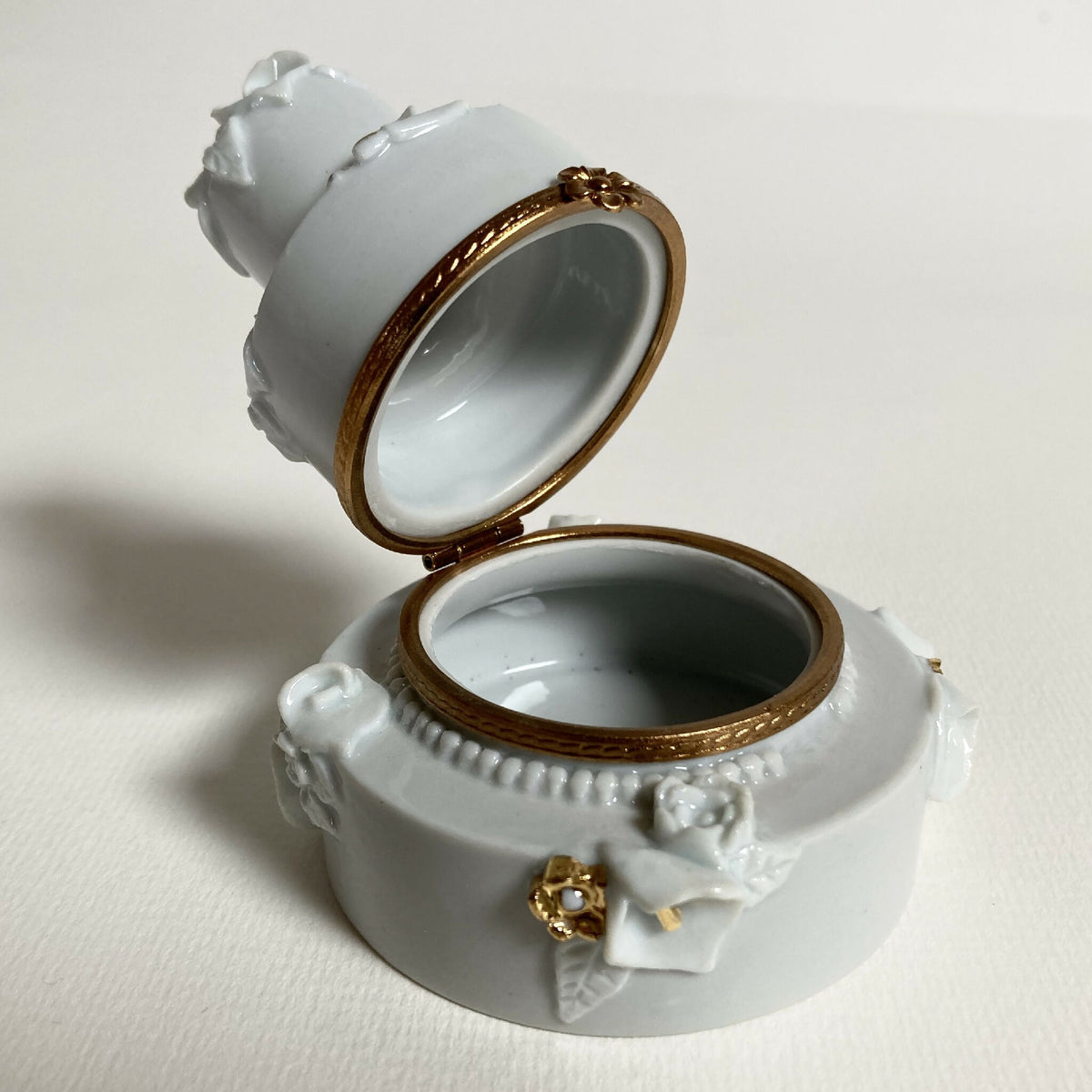 Boîte en porcelaine blanche ouverte, intérieur sombre, bord doré et nœud floral.