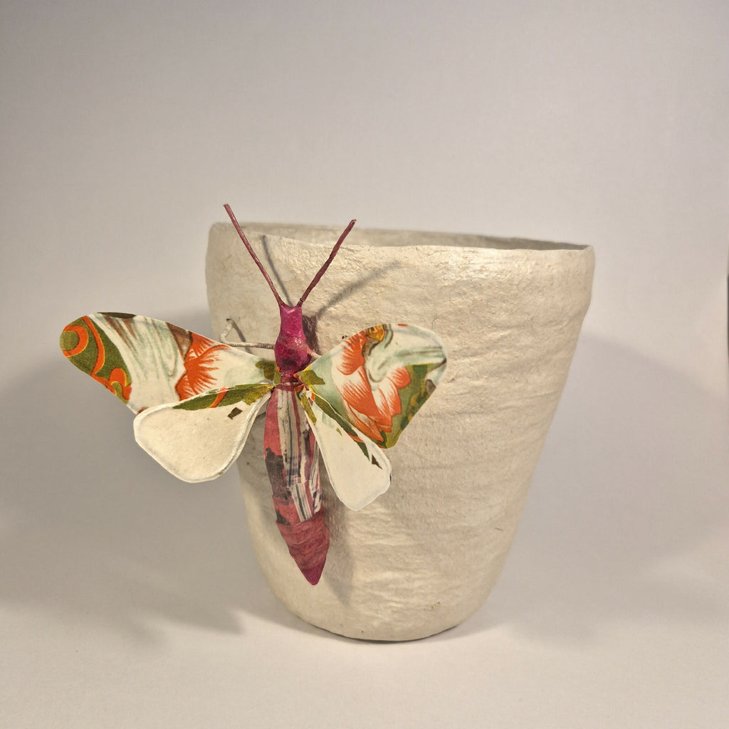 Sculpture de papillon multicolore sur pot en béton clair, vue de trois-quarts