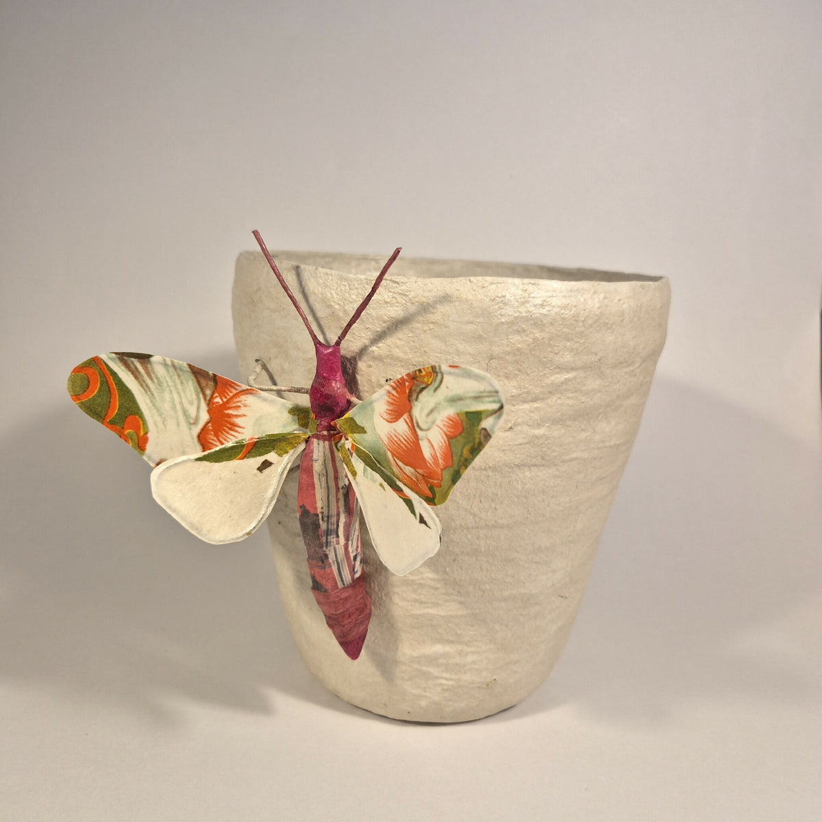 Sculpture de papillon multicolore sur pot en béton clair, vue de trois-quarts