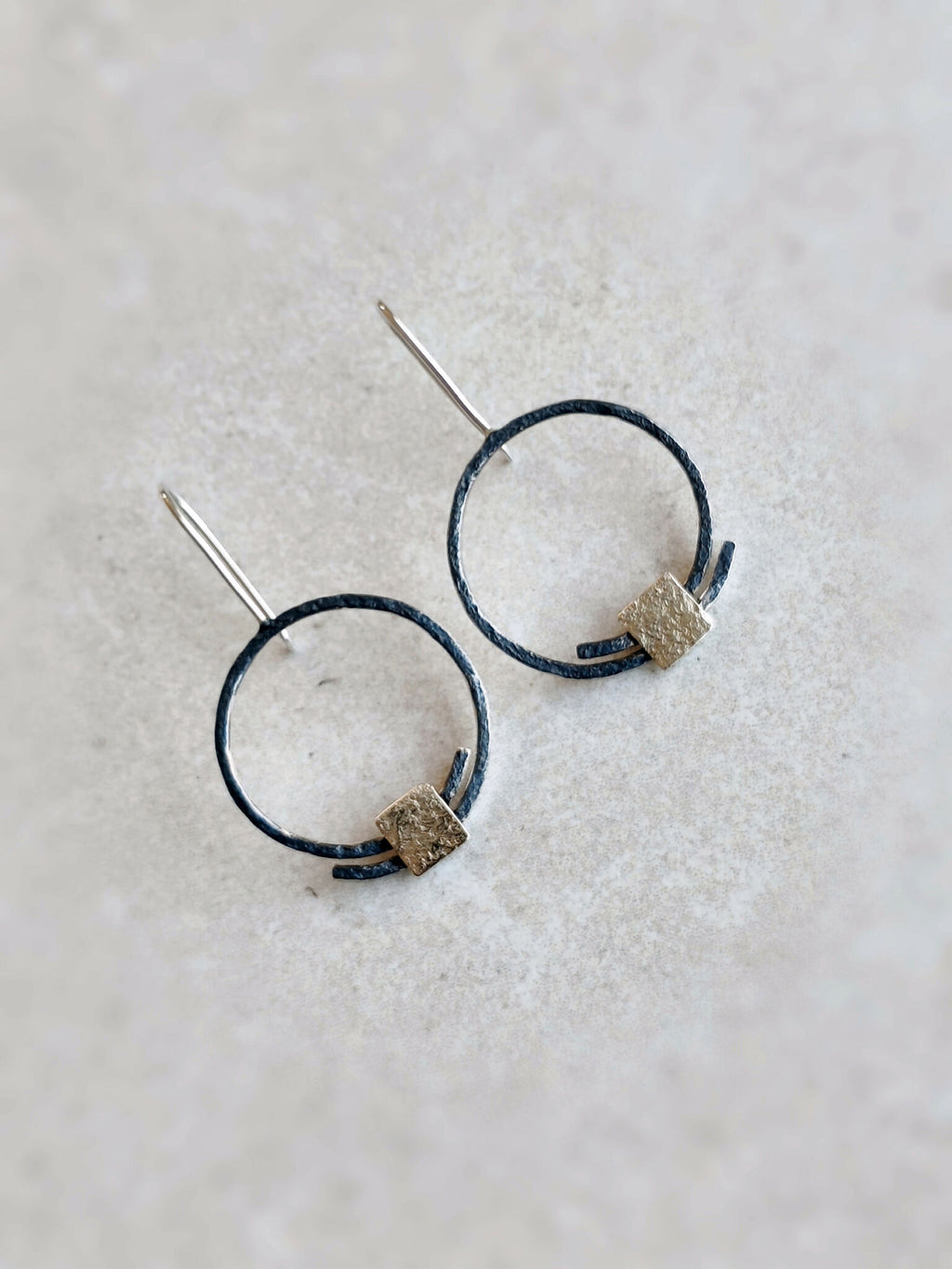 Boucles d’oreilles pendantes : deux anneaux noirs, pastilles carrées dorées texturées, attaches visibles.