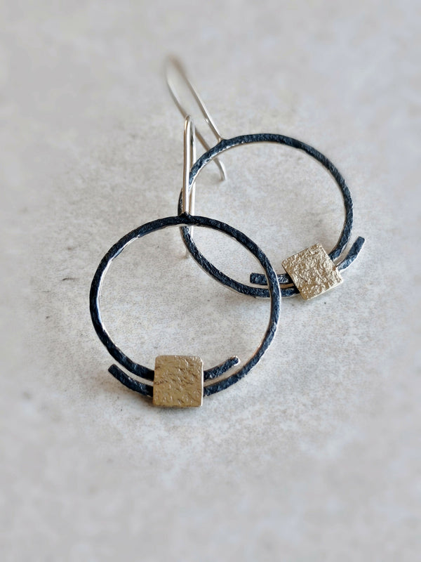 Boucles d’oreilles pendantes : deux anneaux noirs et carrés dorés texturés, tiges argentées.