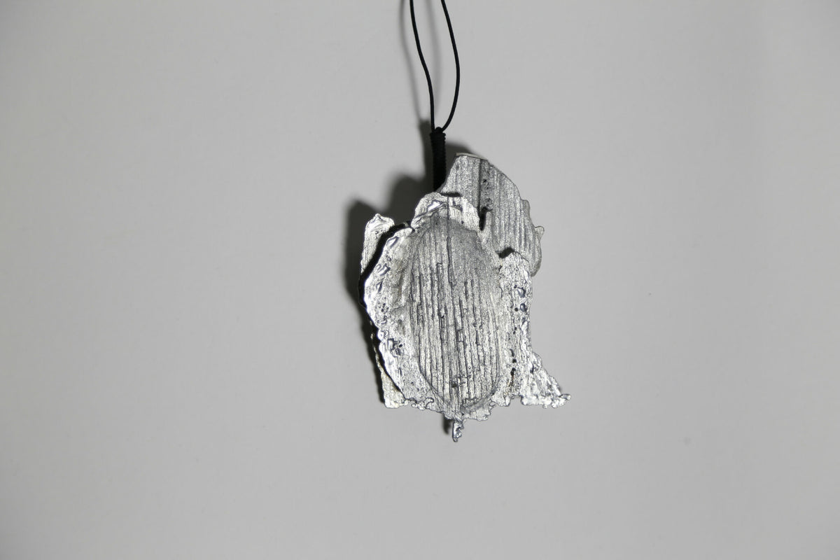 Pendentif argenté-gris en forme de feuille, vue rapprochée et inclinée, cordon noir.