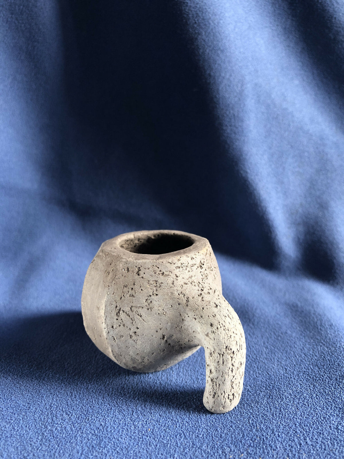 Petit pot en pierre grise avec ouverture circulaire, détails texturés et deux pieds.