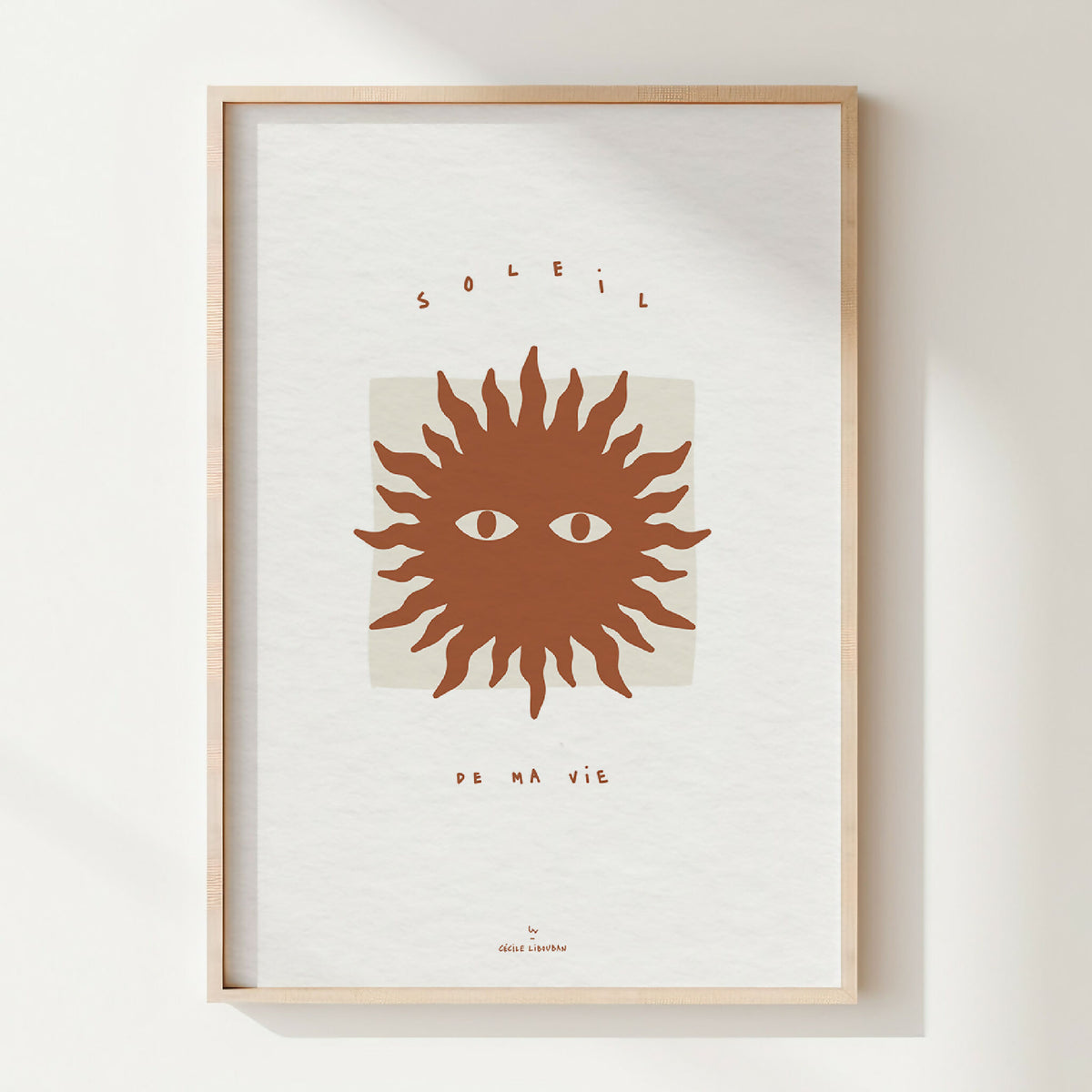 Affiche encadrée : soleil stylisé brun, deux yeux, typographie en haut et bas.