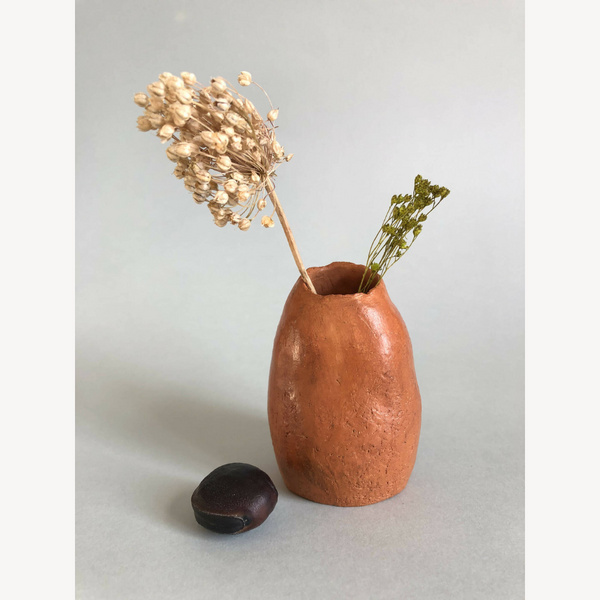 Petit vase en terre cuite orange avec tiges séchées et pierre noire.