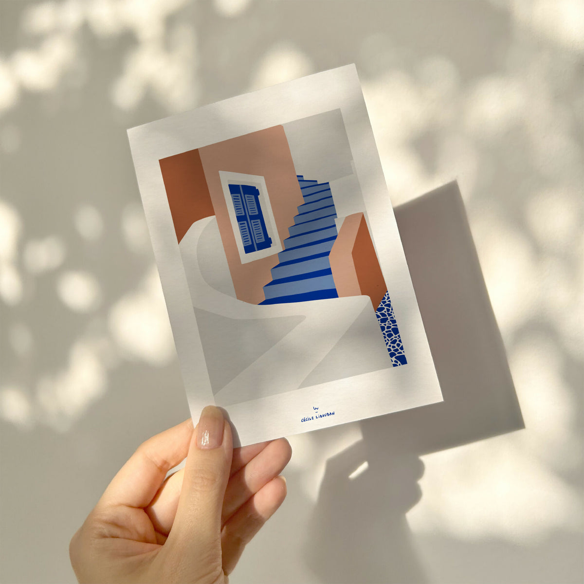 Carte illustrée tenue par une main, escaliers bleus et façade brun/orange