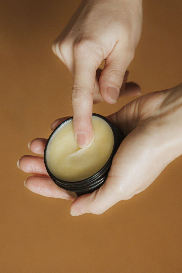 Baume noisette dans un petit pot noir, mains tenant le produit, texture jaune pâle.