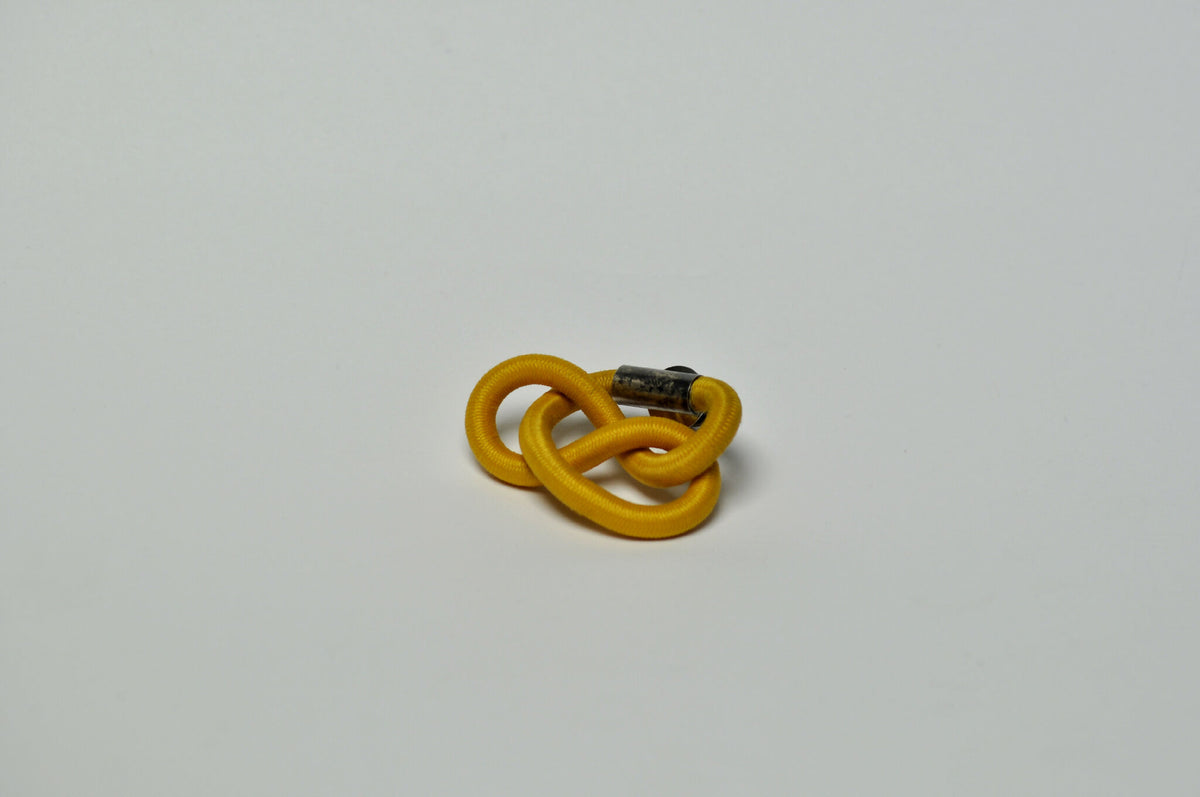 Broche entrelacée jaune, attache métallique visible à l’arrière.