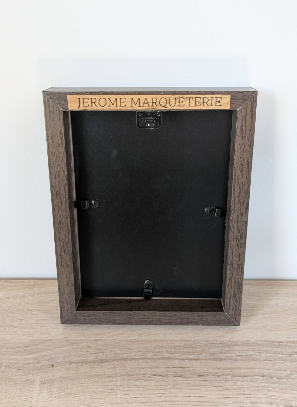 Dos de tableau en bois foncé avec inscription « JEROME MARQUETERIE » et fixations