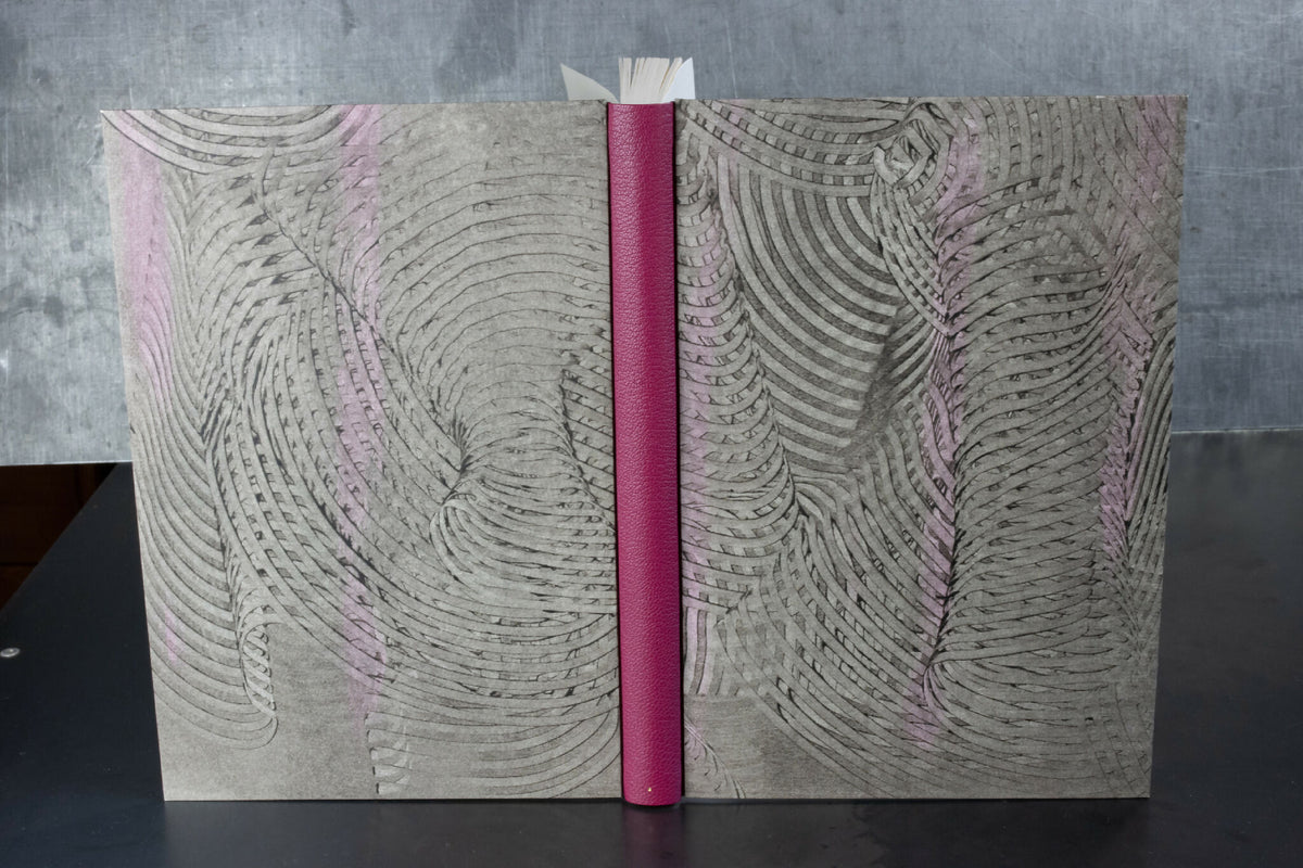 Cahier à couverture cuir gris, motifs ondulés, dos fuchsia et reliure centrale.