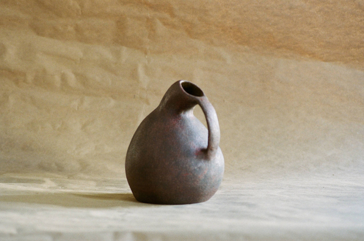 Cruche en céramique sombre, poire arrondie, anse courbée et ouverture supérieure.