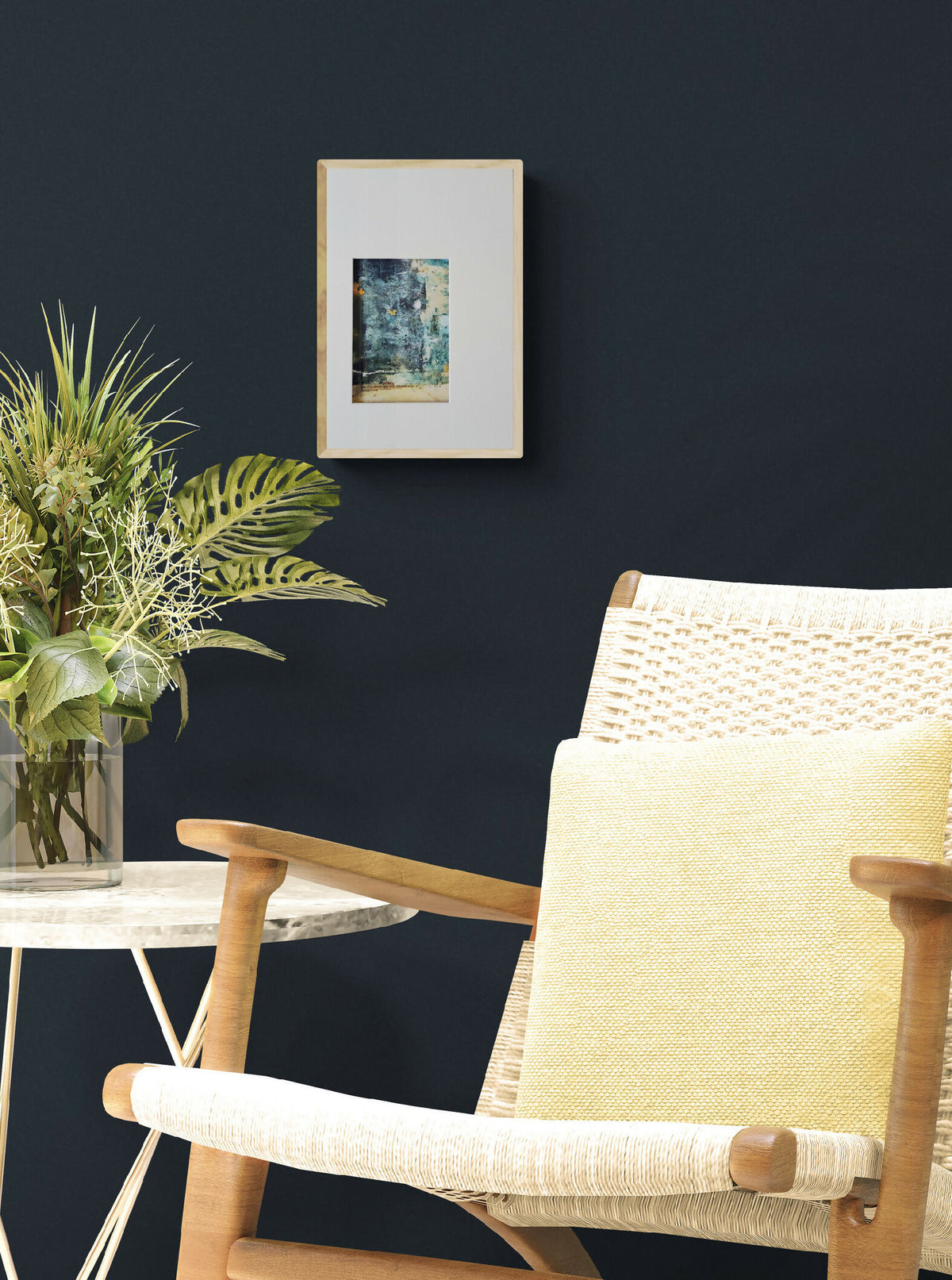 Tableau abstrait dans un cadre beige, motif bleu sur mur sombre, près d’un fauteuil.