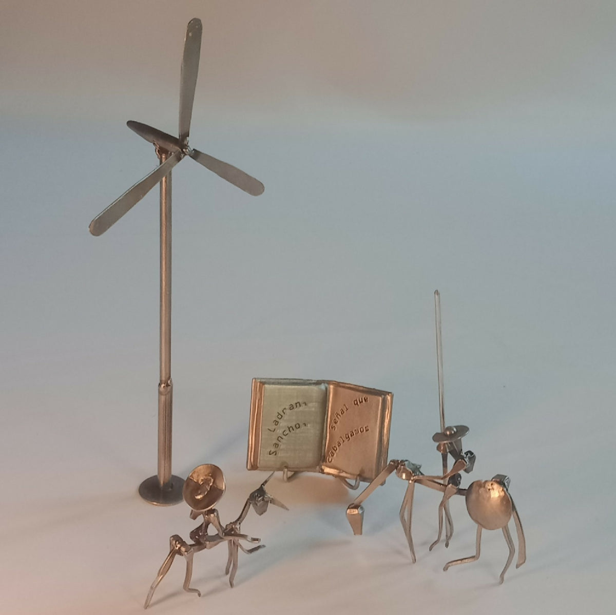 Sculpture en acier : mât à pales, livre ouvert et petits personnages sur socle.