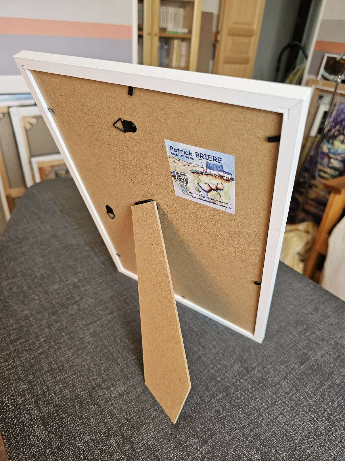 Dos de tableau encadré en carton, étiquette et attache murale visibles.