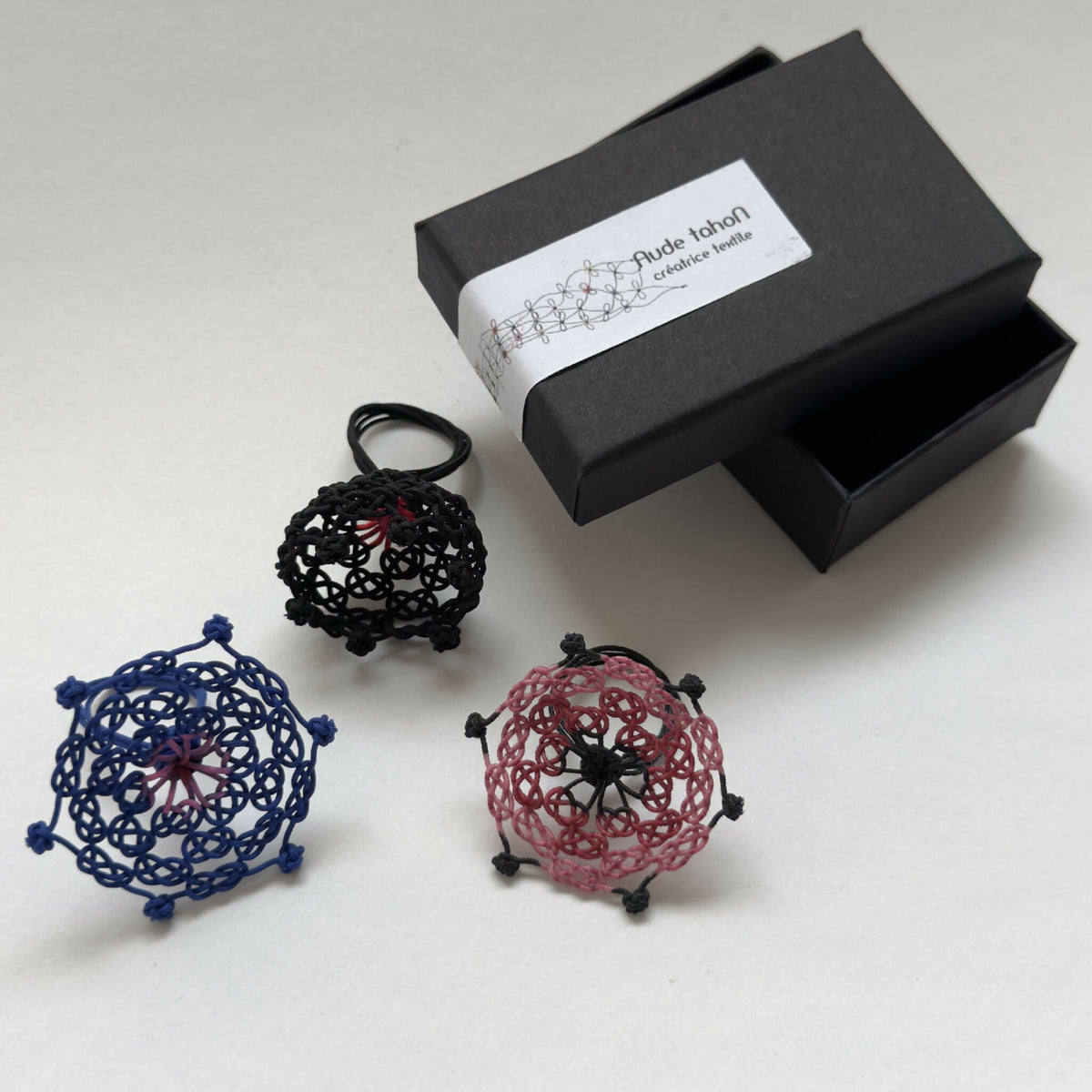 Bague en macramé bleue en forme de trèfle avec centre rose, posée sur des boîtes