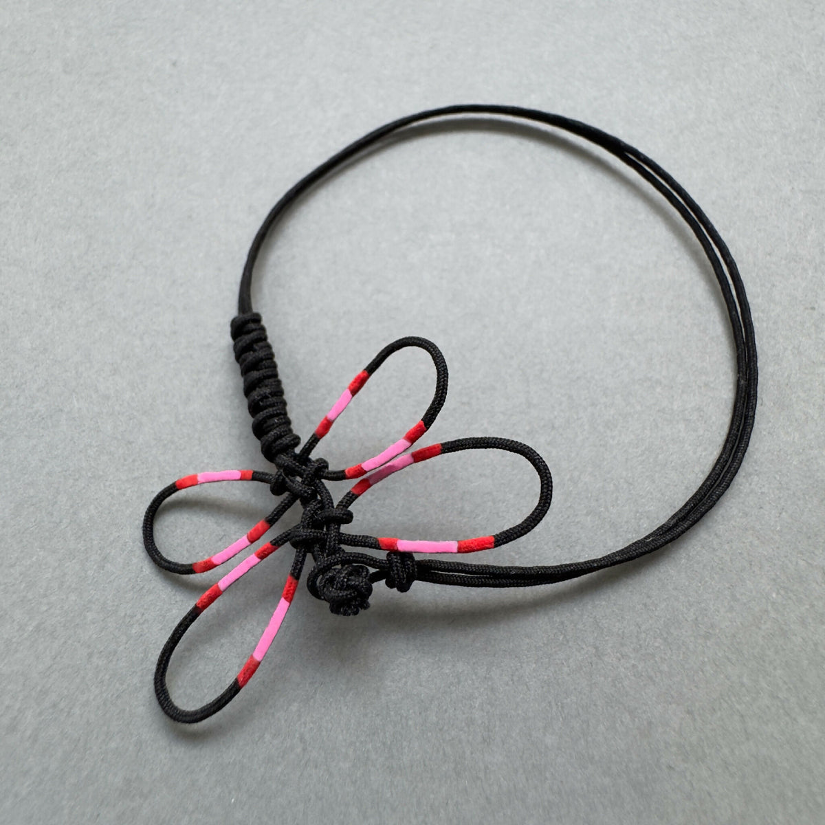Bracelet noir en cordon tressé avec libellule rose et rouge, nœud central visible.