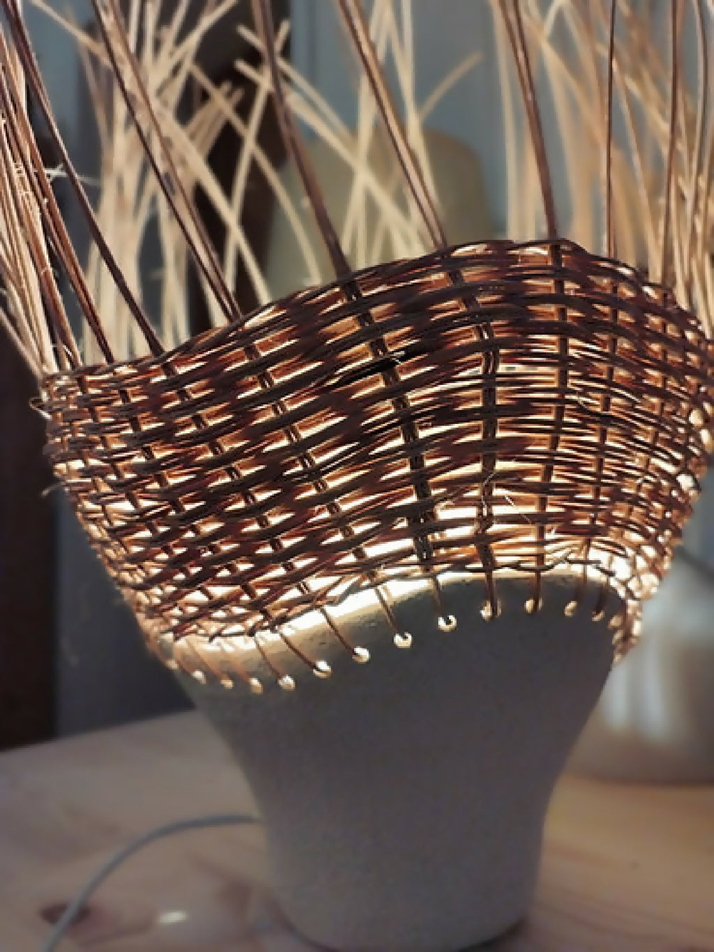 Luminaire en rotin brun tressé sur pot en grès clair, bord ajouré illuminé.