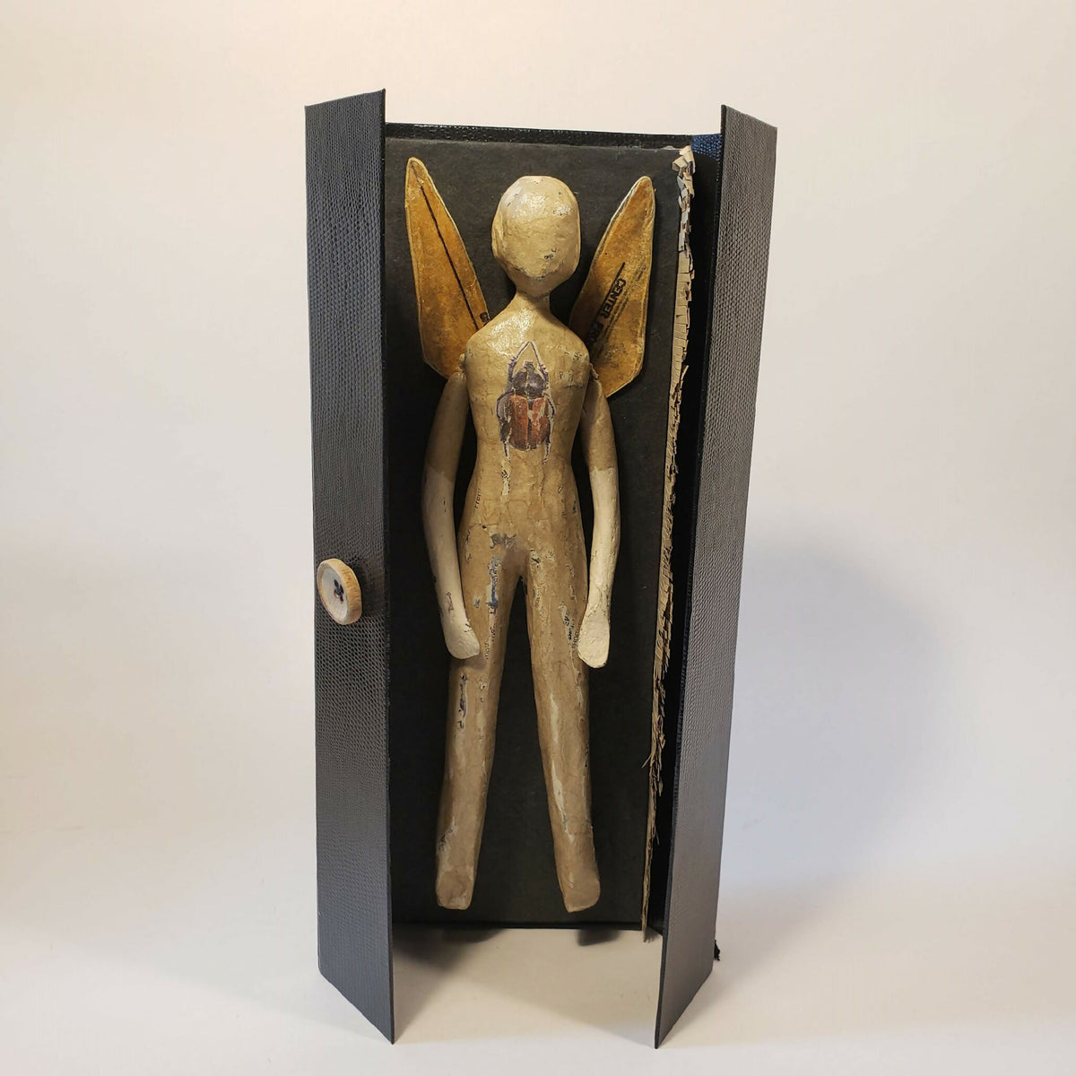 Sculpture papier de poupée beige aux ailes dorées, dans un écrin noir ouvert