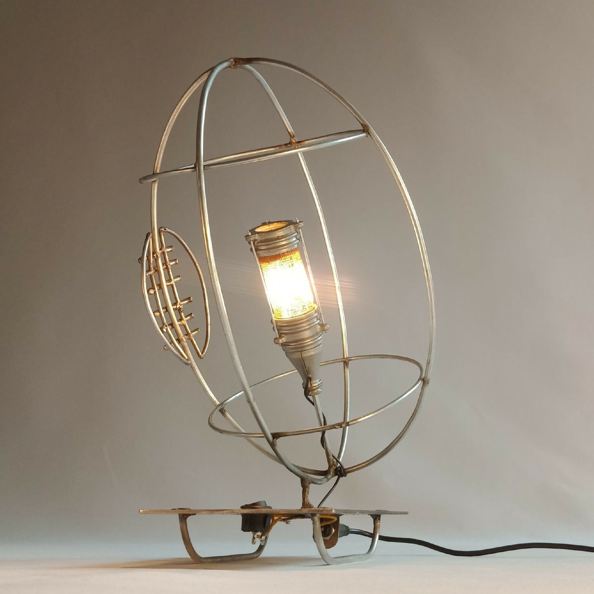 Lampe RUGBY allumée : cage ronde dorée, ampoule éclairante et base sur pied