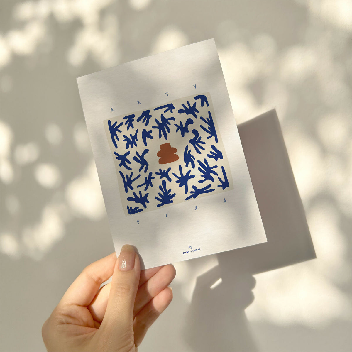 Carte illustrée bleue, motif végétal et élément terracotta au centre, tenue par une main