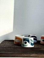 Petites tasses trapèzes blanches à motifs bleus, verts et orange, alignées sur tablette.