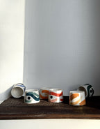 Plusieurs petites tasses trapèzes blanches à motifs bleus, verts et orange, sur tablette.