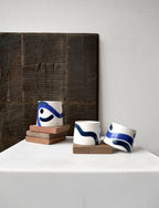 Petite tasse trapèze blanche à motifs bleus, vue en perspective sur socle.