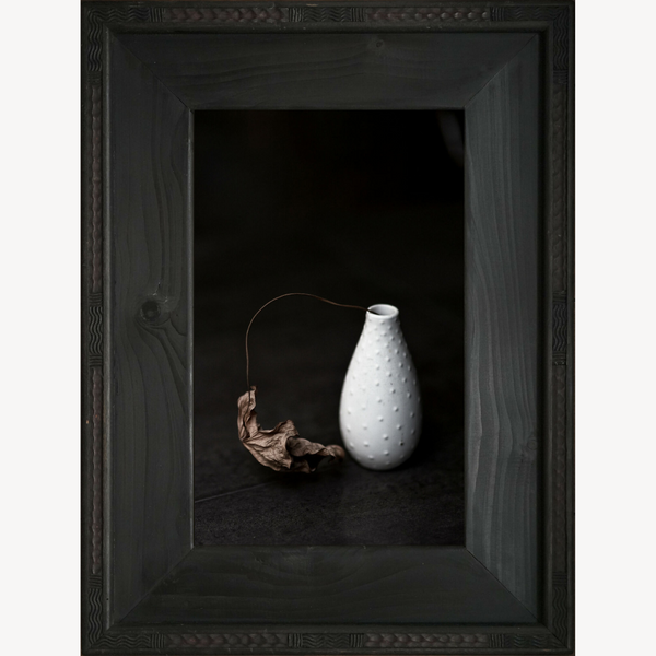 Cadre avec vase blanc texturé et branche de feuille morte séchée.