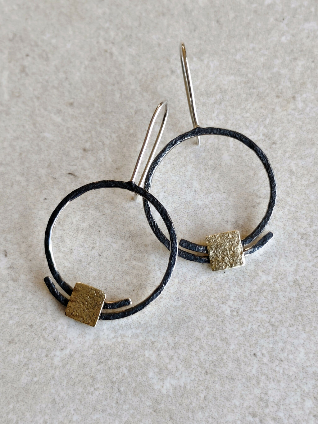Boucles d’oreilles pendantes en anneaux : deux anneaux noirs avec carrés dorés texturés, tiges fines.