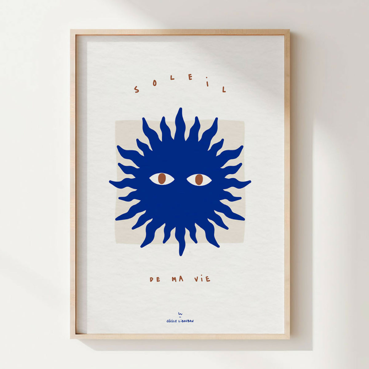 Affiche encadrée : soleil stylisé bleu, deux yeux, typographie en haut et bas.