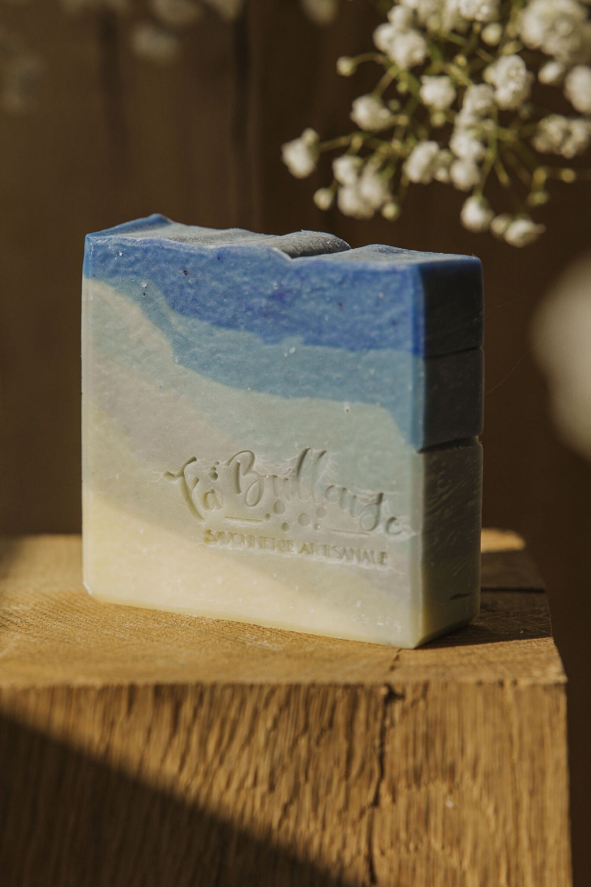 Savon rectangulaire bleu et beige, face avant avec logo gravé, près de fleurs