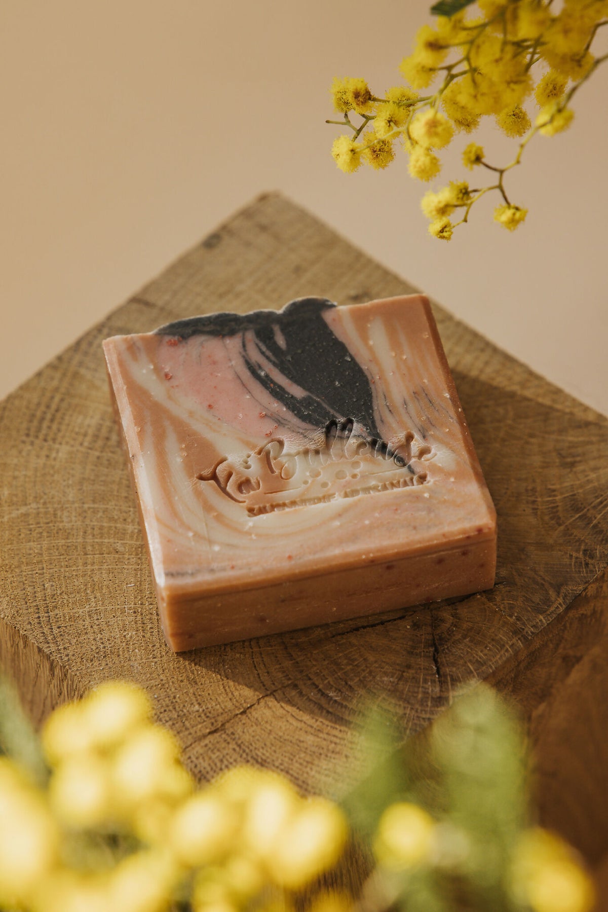 Savon en barre marbrée rosé et noir, sur toile de jute, près de fleurs jaunes.