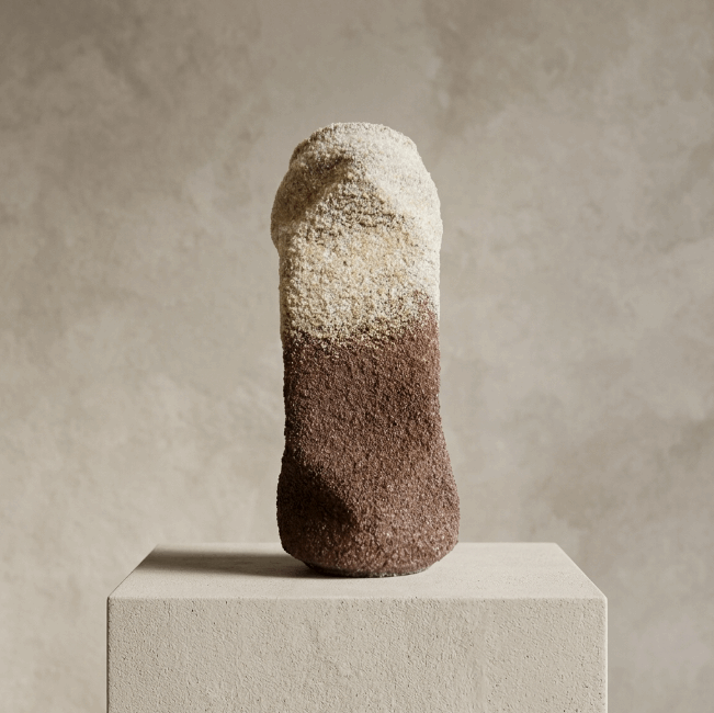 Sculpture cylindrique granuleuse, haut beige clair, bas brun foncé, posée sur socle.