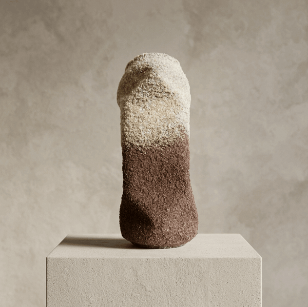 Sculpture cylindrique granuleuse, haut beige clair, bas brun foncé, posée sur socle.