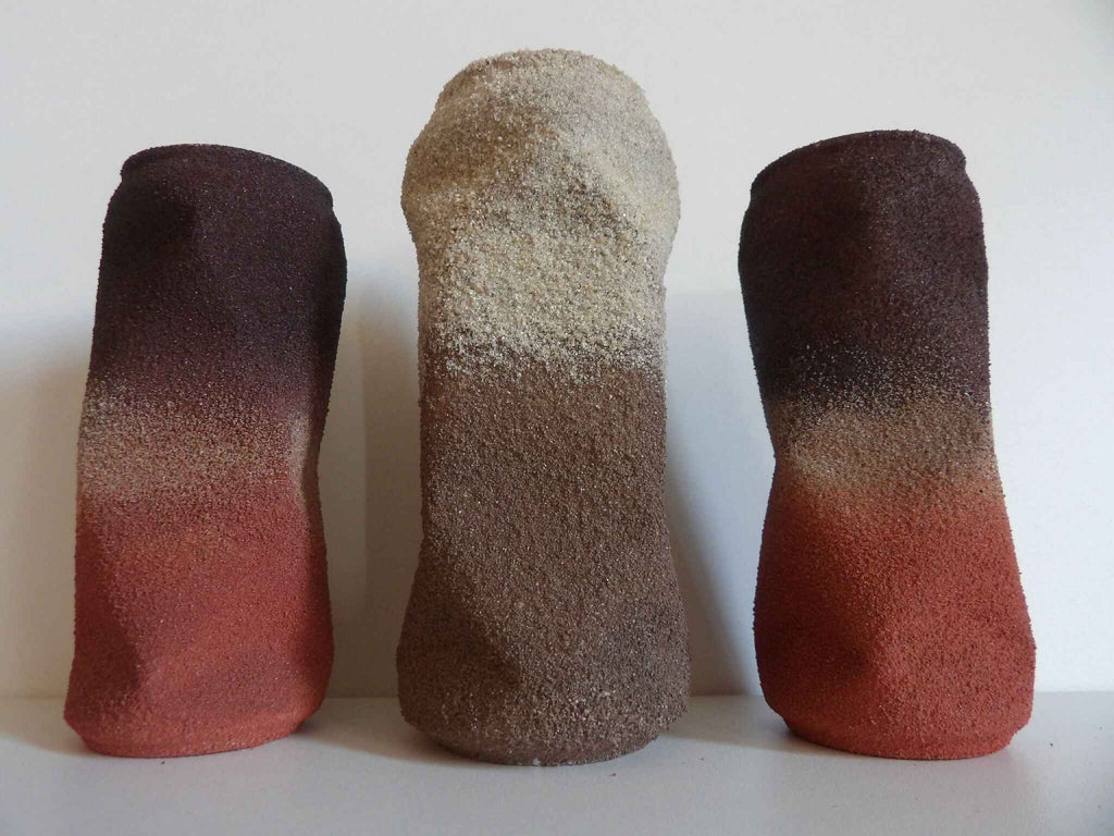 Sculptures cylindriques en trio : deux foncées et une claire, textures granuleuses.