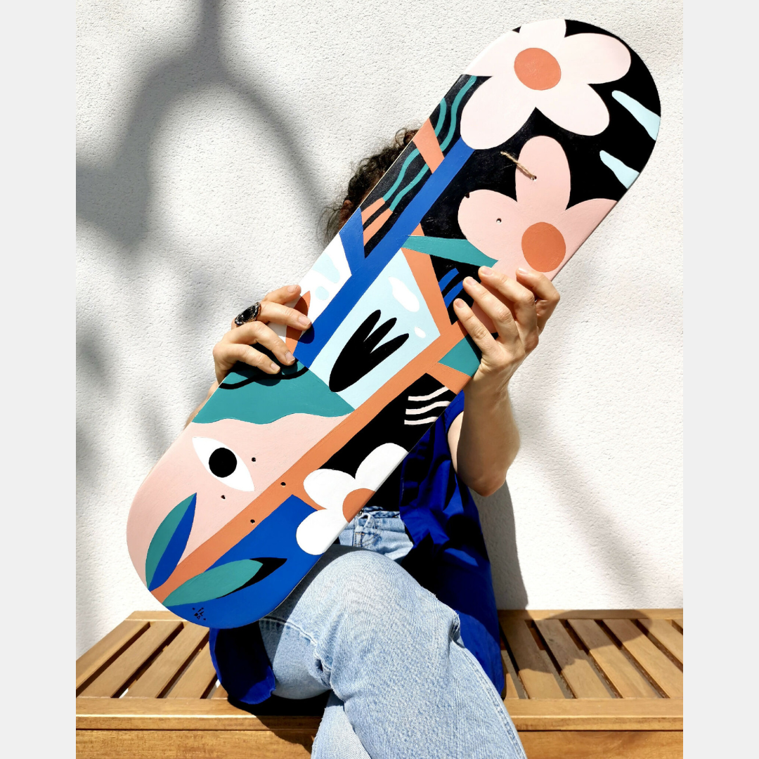 Skateboard illustré, motifs de visages et fleurs, couleurs vives bleu, orange et noir.
