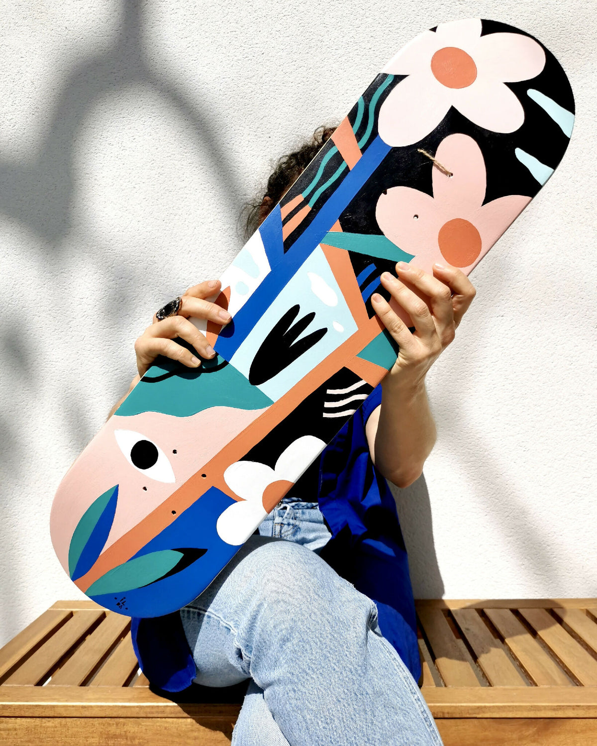 Skateboard à motifs de visages et fleurs, couleur bleu et noir, tenu à deux mains.
