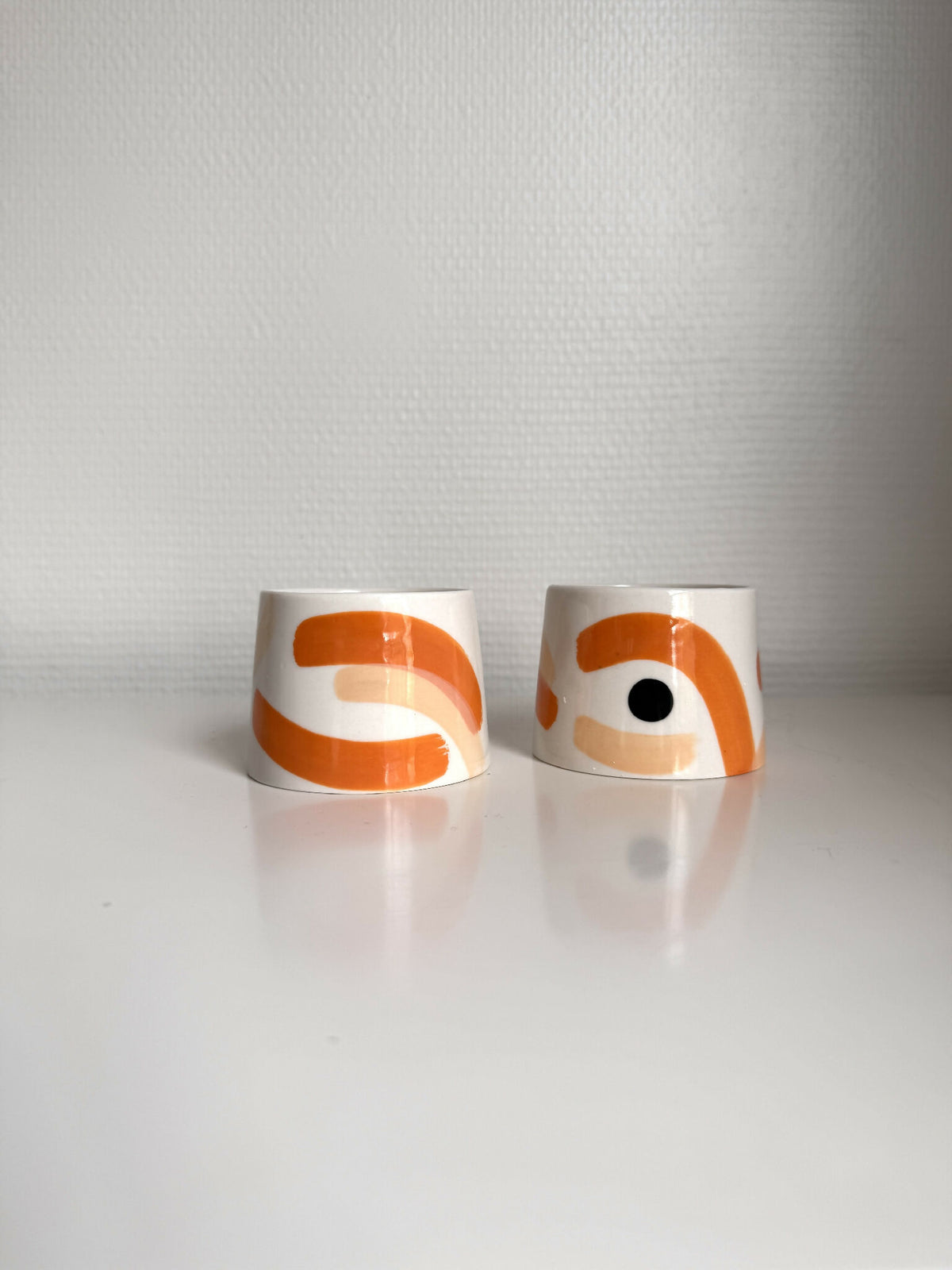 Petites tasses trapèzes blanches à motifs orange, deux tasses avec reflet au sol.