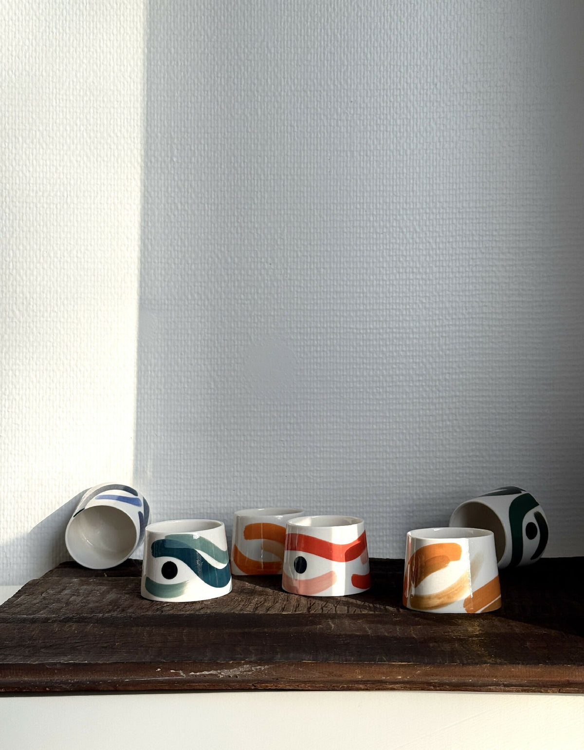 Plusieurs petites tasses trapèzes blanches à motifs bleus, verts et orange, sur tablette.