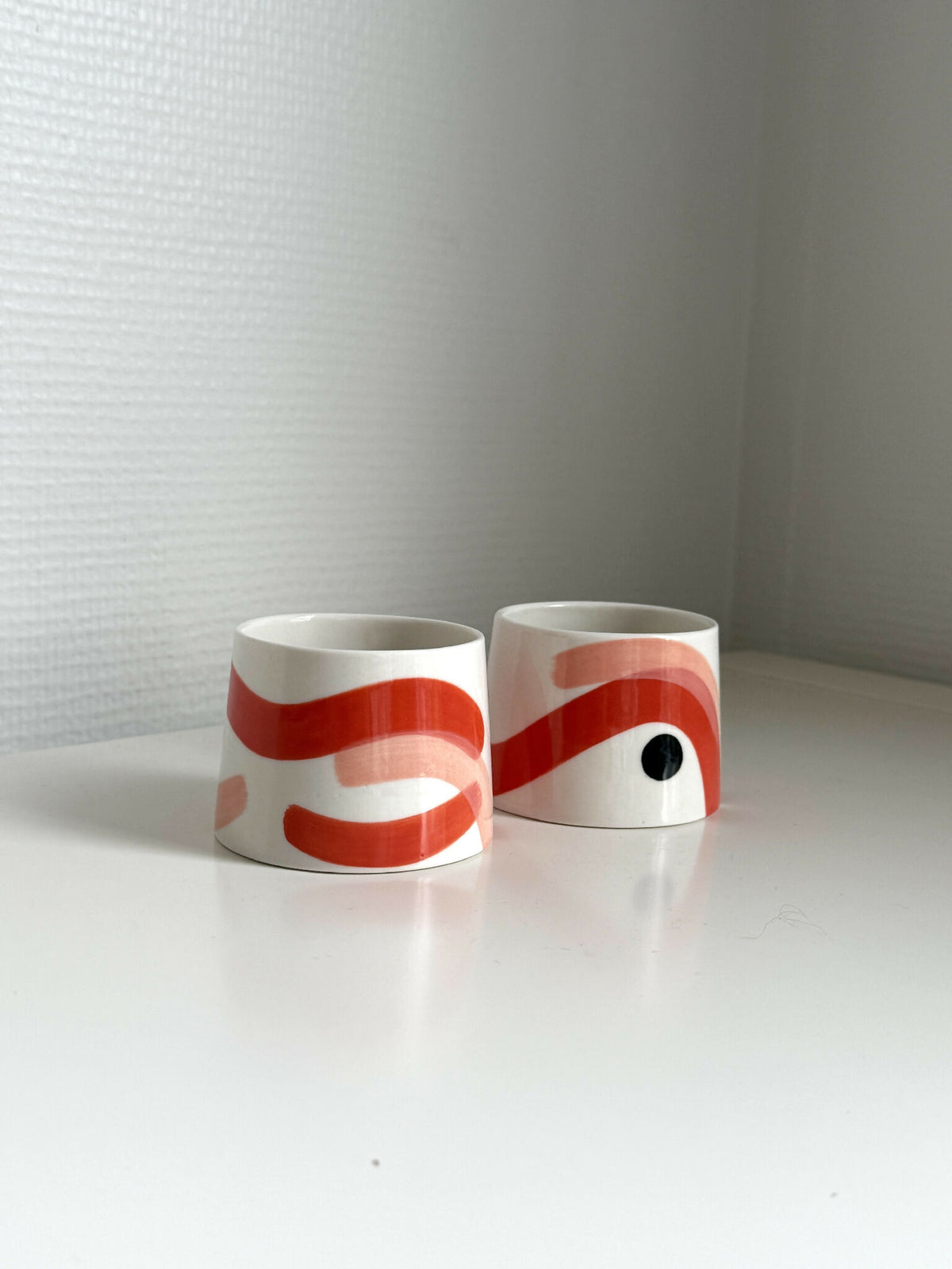 Petites tasses trapèzes blanches à motifs rouges et yeux noirs, deux exemplaires.